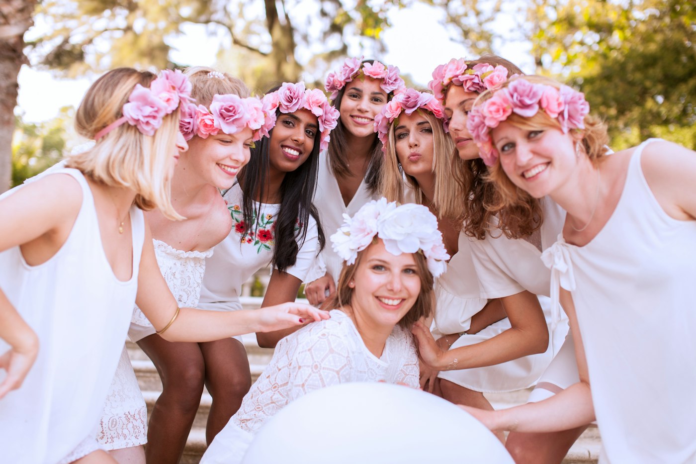 Die Braut mit weißem Blumenkranz sitzt vor sieben Freundinnen mit rosa Rosenkränzen im Haar, alle lachen draußen in die Kamera