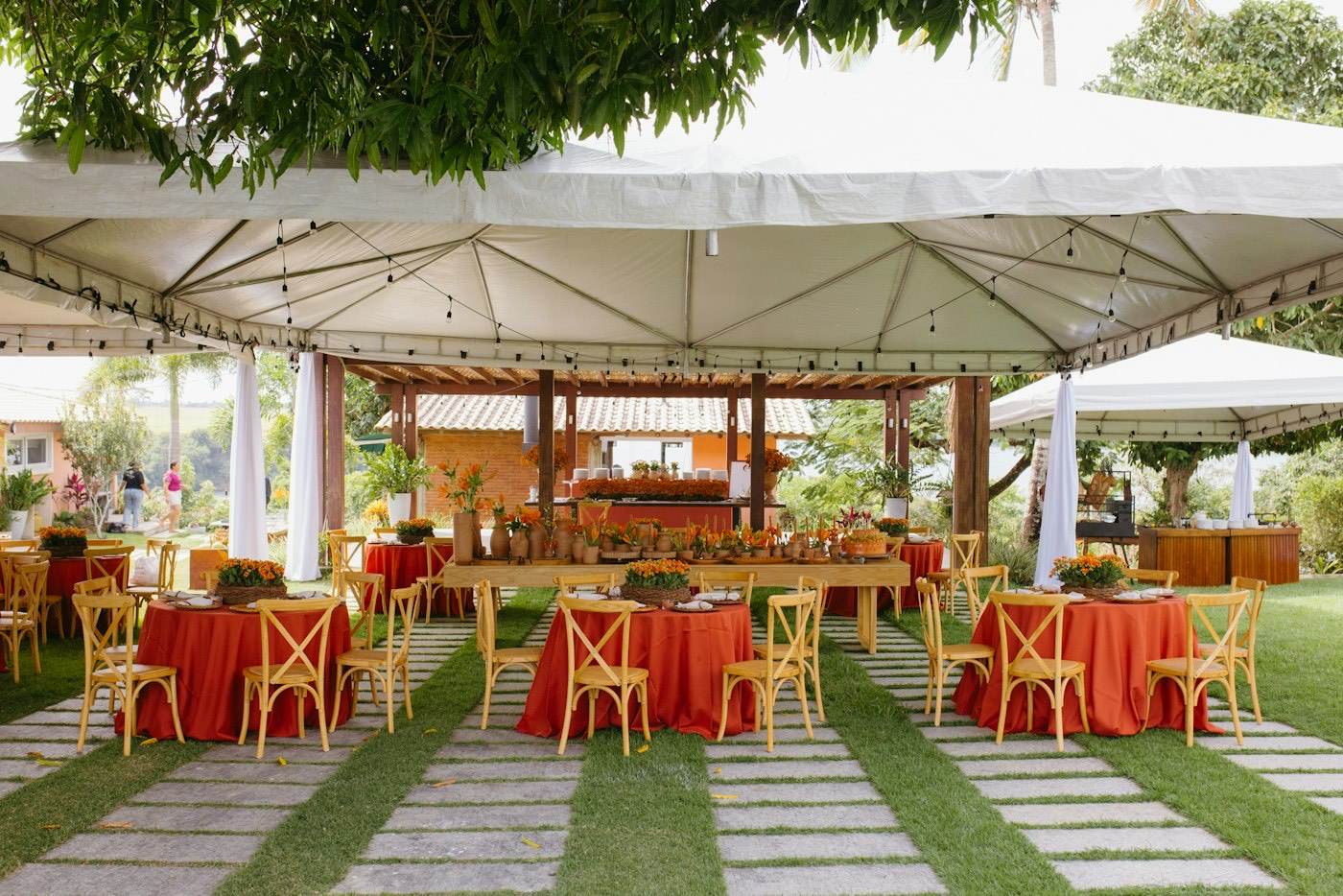 Ein weißes Mietzelt auf dem Rasen mit runden Tischen in orangefarbenen Decken und Holzstühlen für eine Gartenhochzeit