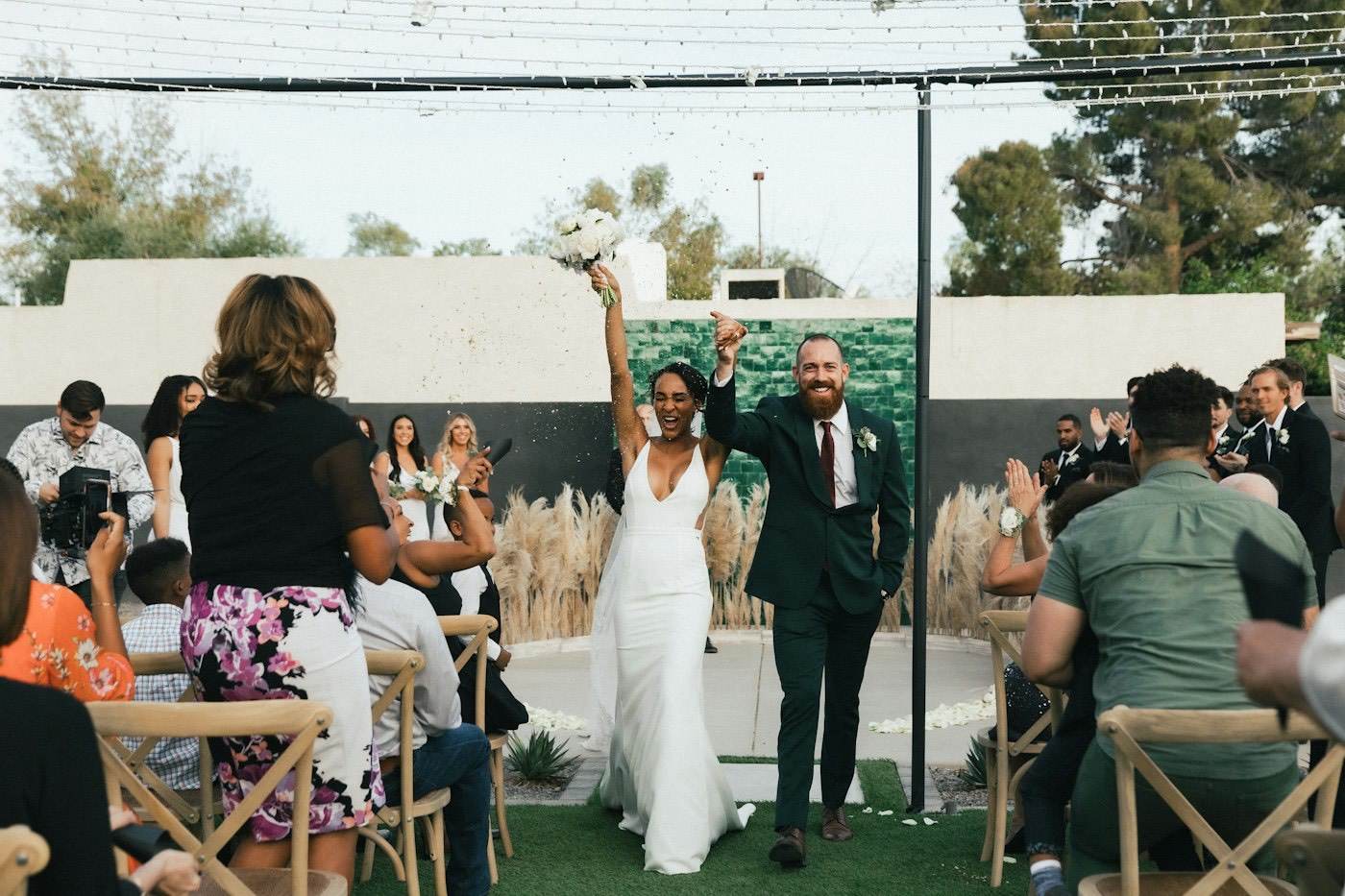 Newlyweds raise their hands as they walk back down the aisle past guests, with pampas grass behind the ceremony spot