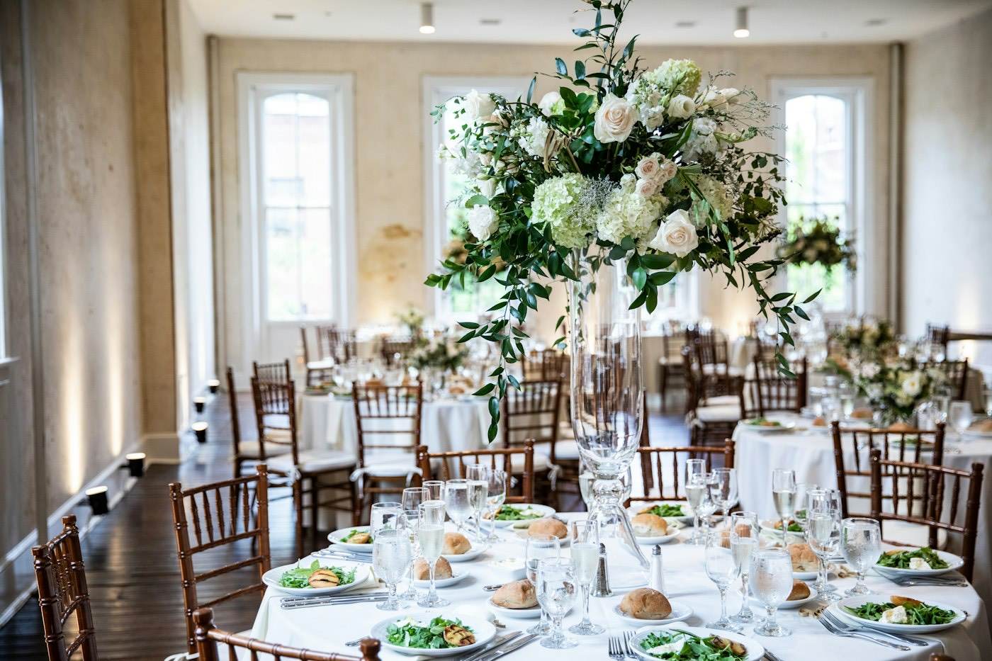 A tall white flower centerpiece on a glass stand on one table, with low arrangements on the tables behind it