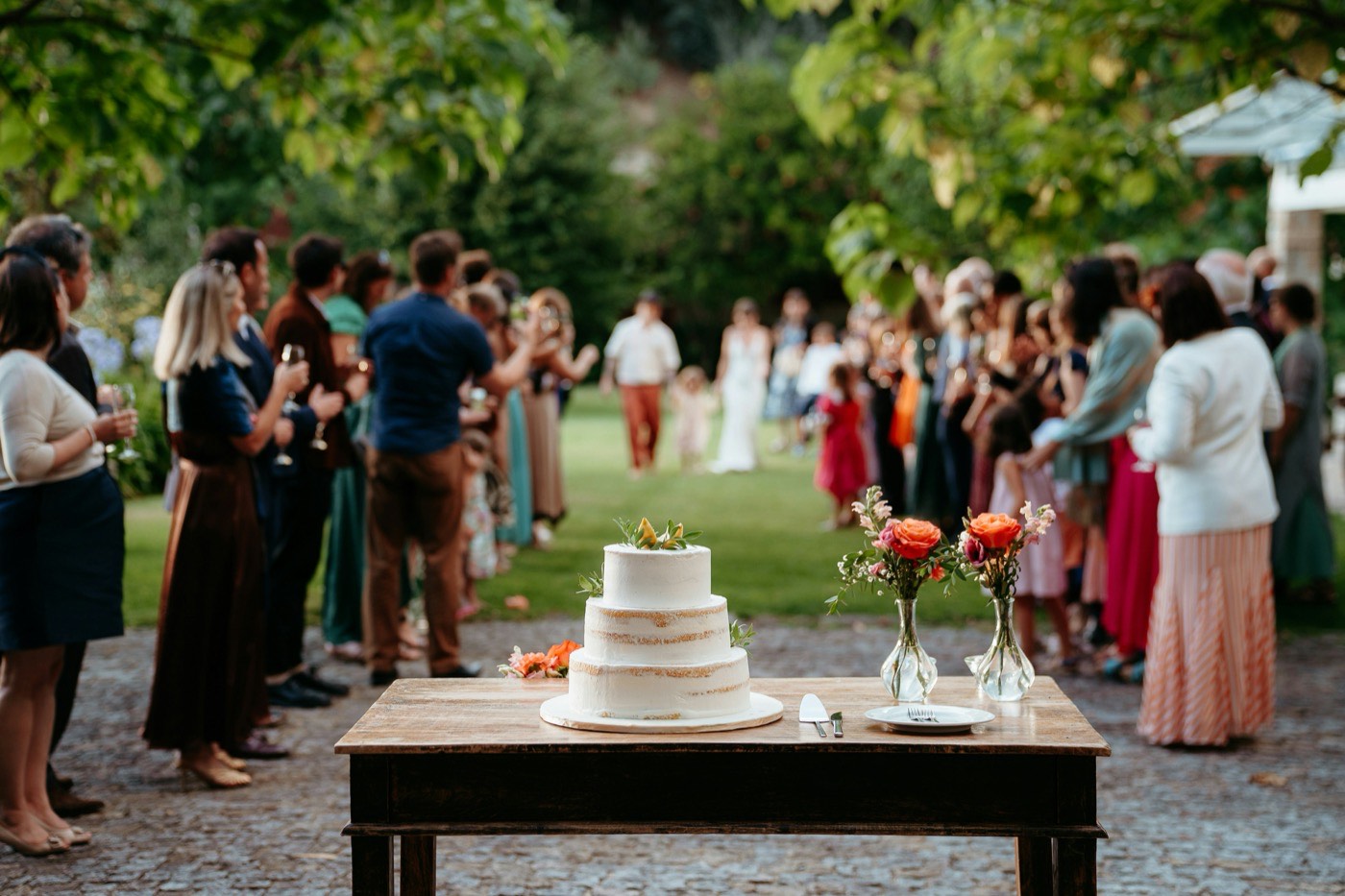 Eine dreistöckige Hochzeitstorte auf einem Holztisch neben zwei kleinen Vasen mit orangefarbenen Rosen, dahinter Gäste auf dem Rasen