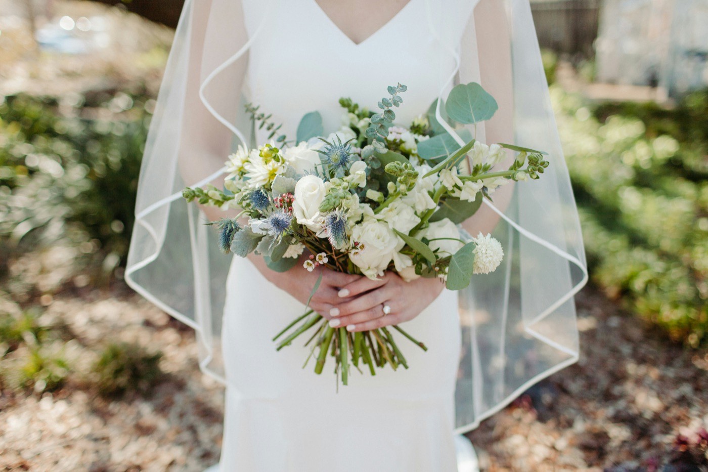 Eine Braut in weißem Kleid und Schleier hält einen lockeren Strauß aus weißen Rosen, Disteln und Eukalyptus im Garten