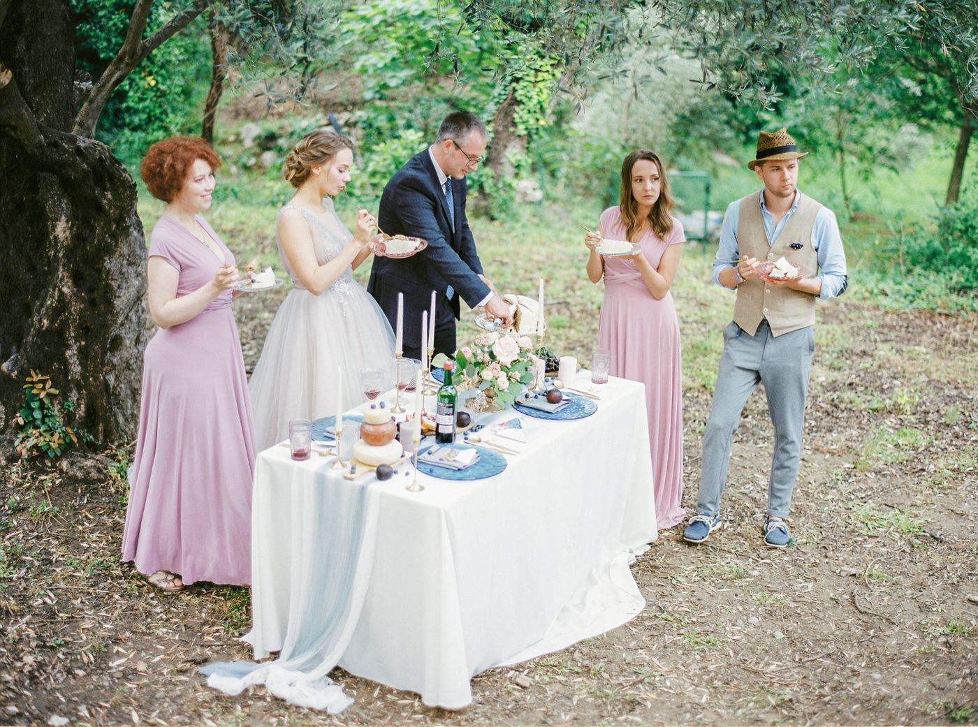 Los novios y sus amigos comen tarta en platitos alrededor de una mesa al aire libre bajo los olivos