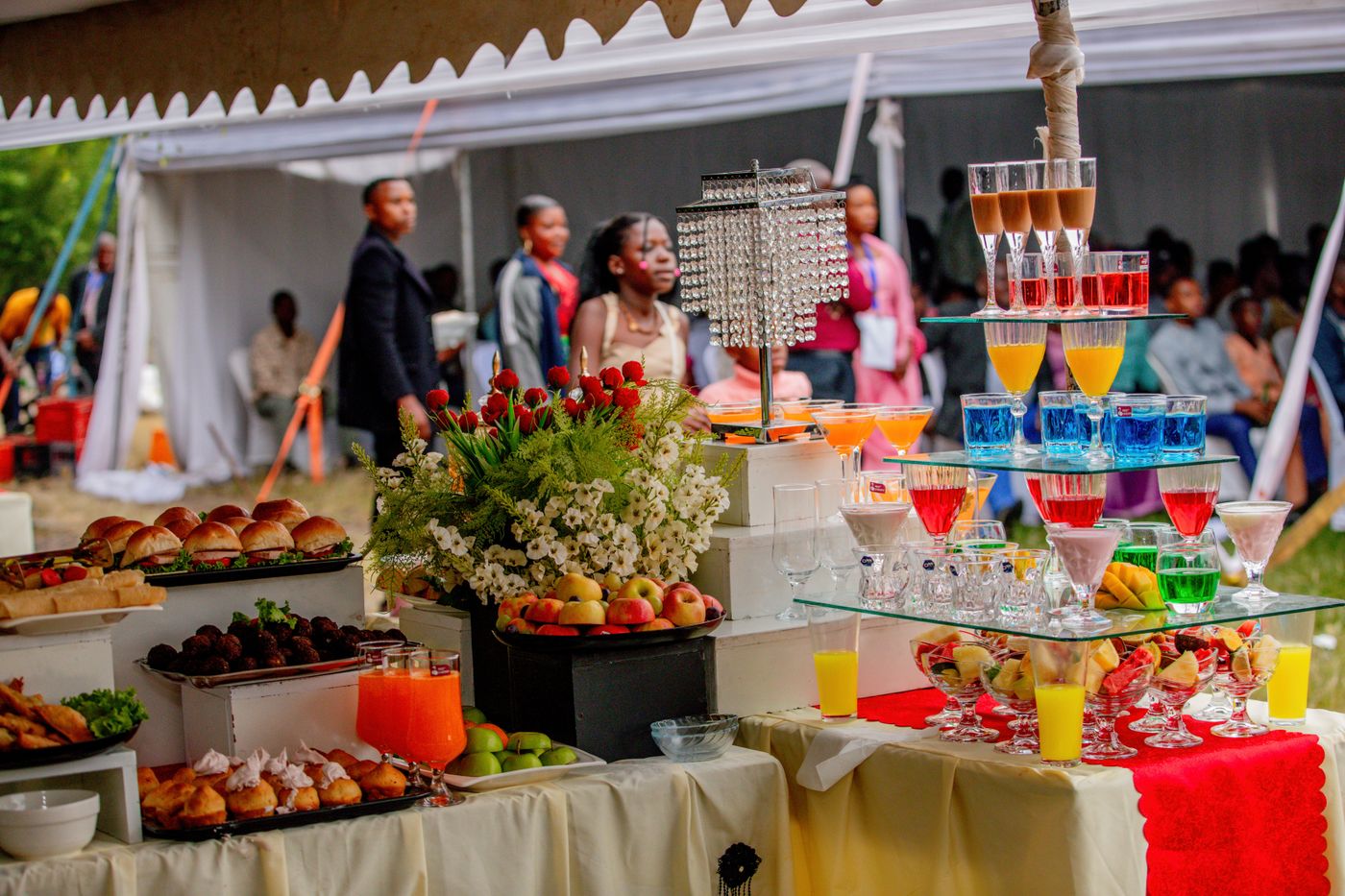 Mesa de bufé en una boda bajo carpa con mini hamburguesas, fruta y vasos de colores mientras los invitados charlan al fondo