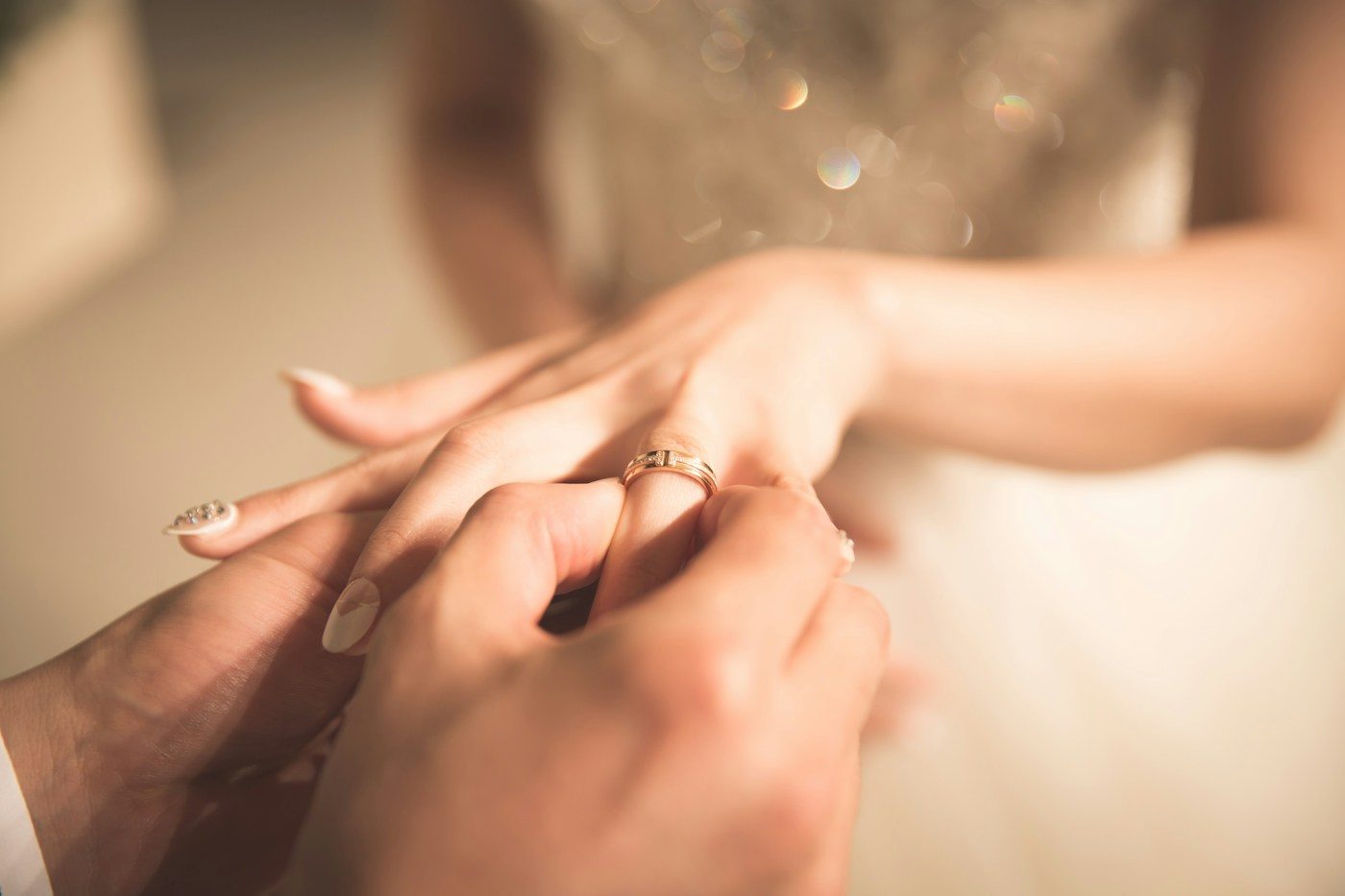 Der Bräutigam steckt der Braut einen schmalen Goldring an, im Hintergrund ihr glitzerndes Brautkleid