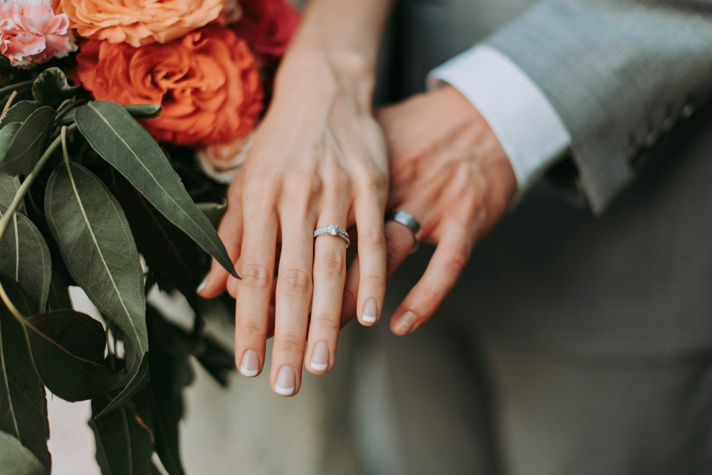Braut und Bräutigam legen die Hände nebeneinander, sie mit schmalem Silberring, er mit dunkelgrauem Ring, daneben ein Strauß orangefarbener Rosen