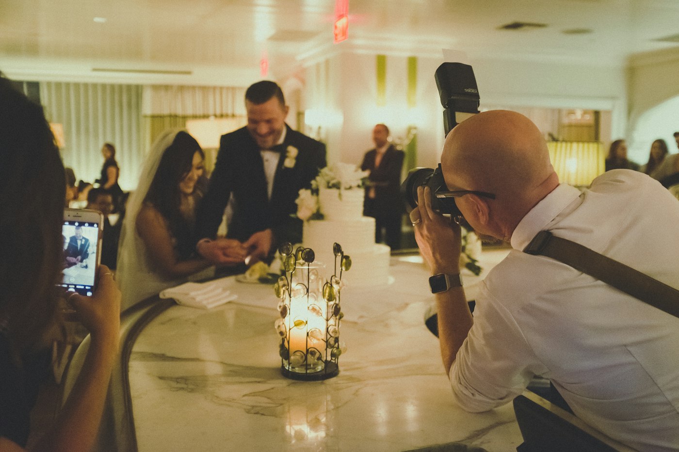A bride and groom laugh as they cut a white tiered cake in a restaurant dining room while a photographer and a guest with a phone capture it