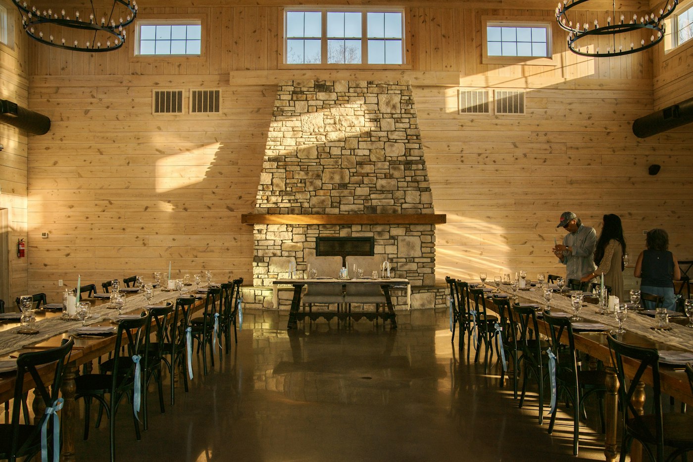 Long wooden tables with gauze runners, candles and glasses in a plain wood-paneled hall with a stone fireplace while guests arrive