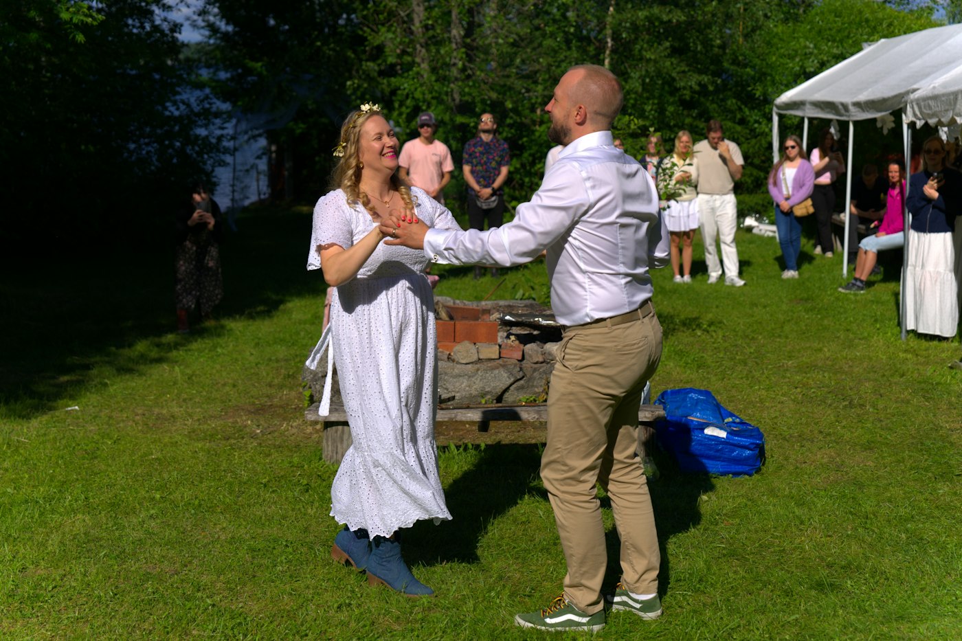 A bride and groom dance on the grass at a casual lakeside park party while guests watch from beside a white pop-up tent