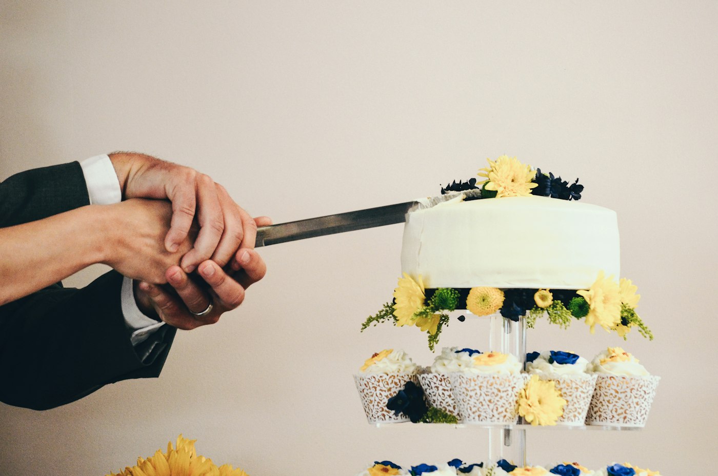 A couple holds a cake knife together over a small white cake with yellow flowers, sitting on a tower of cupcakes