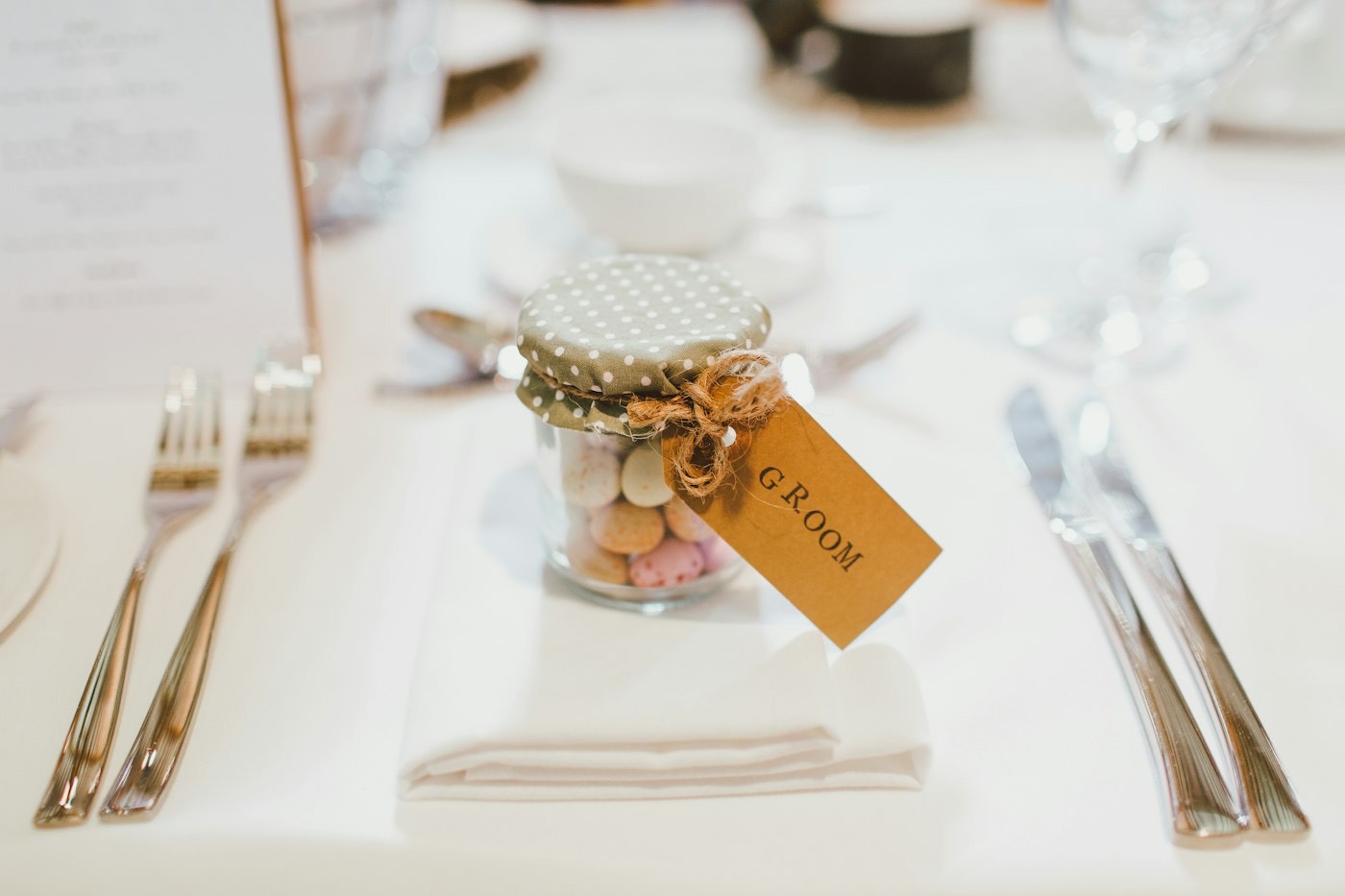 A small jar of sugared almonds with a kraft name tag sits on a napkin at a wedding place setting