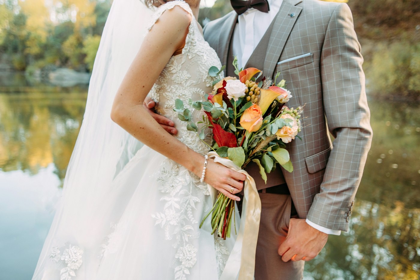Close-up of a fall bridal bouquet with orange calla lilies, roses and eucalyptus held against the groom’s tweed suit