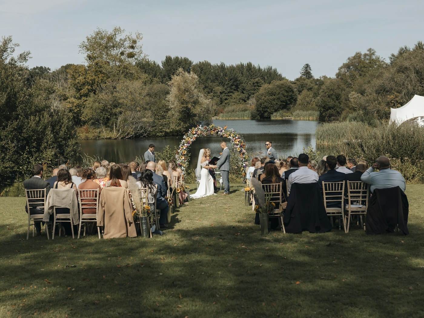 Guests seated on a lawn watch an outdoor ceremony under a flower arch beside a lake in early autumn