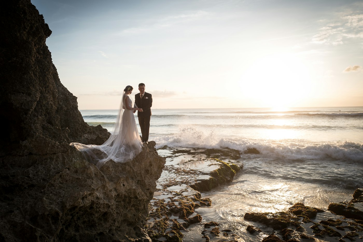 Una novia con un velo largo y un novio con traje oscuro se cogen de la mano sobre una roca junto al mar al atardecer