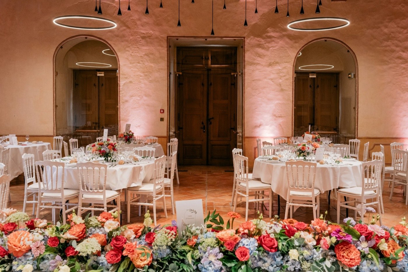 Runde Hochzeitstische mit je einem Blumengesteck, davor eine lange, niedrige Reihe aus roten, orangen und blauen Blumen