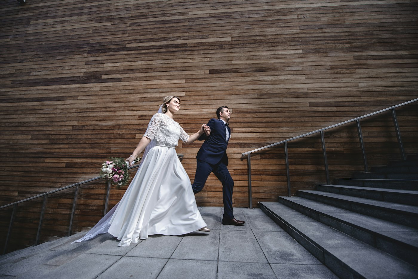 Una novia con top de encaje y falda de satén y un novio con traje azul marino caminan de la mano junto a una pared moderna de madera y unas escaleras de piedra