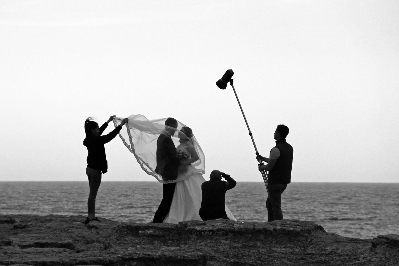 Schwarz-Weiß-Foto eines Hochzeitsshootings auf einer Klippe am Meer: Eine Assistentin hebt den Schleier der Braut, ein Fotograf kniet, ein anderer hält eine Leuchte an einer Stange