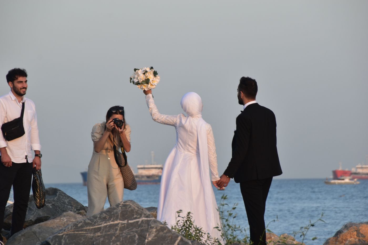 Eine Fotografin hockt auf den Felsen am Meer und fotografiert eine Braut, die ihren Strauß hochhält und den Bräutigam an der Hand hält