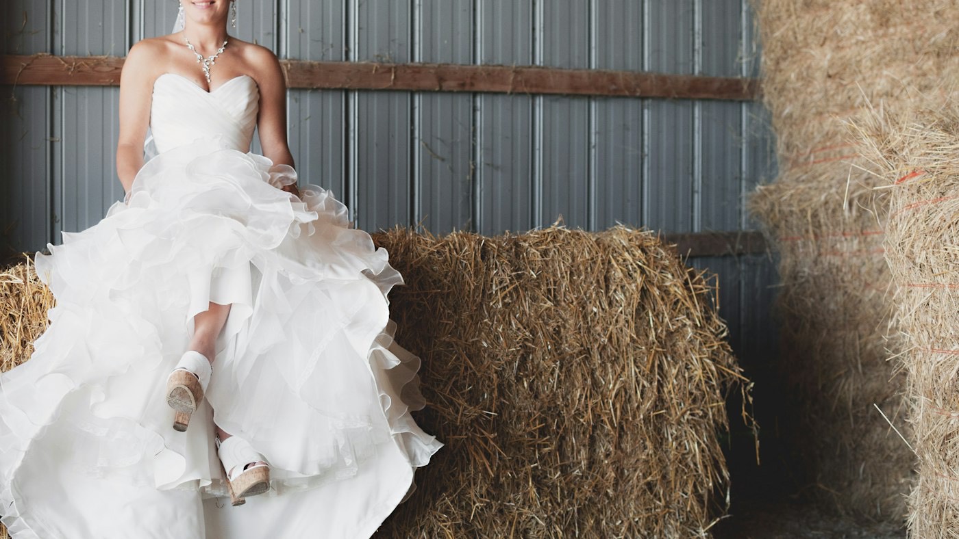Une mariée en robe à volants assise sur une botte de paille dans une grange aux murs de tôle