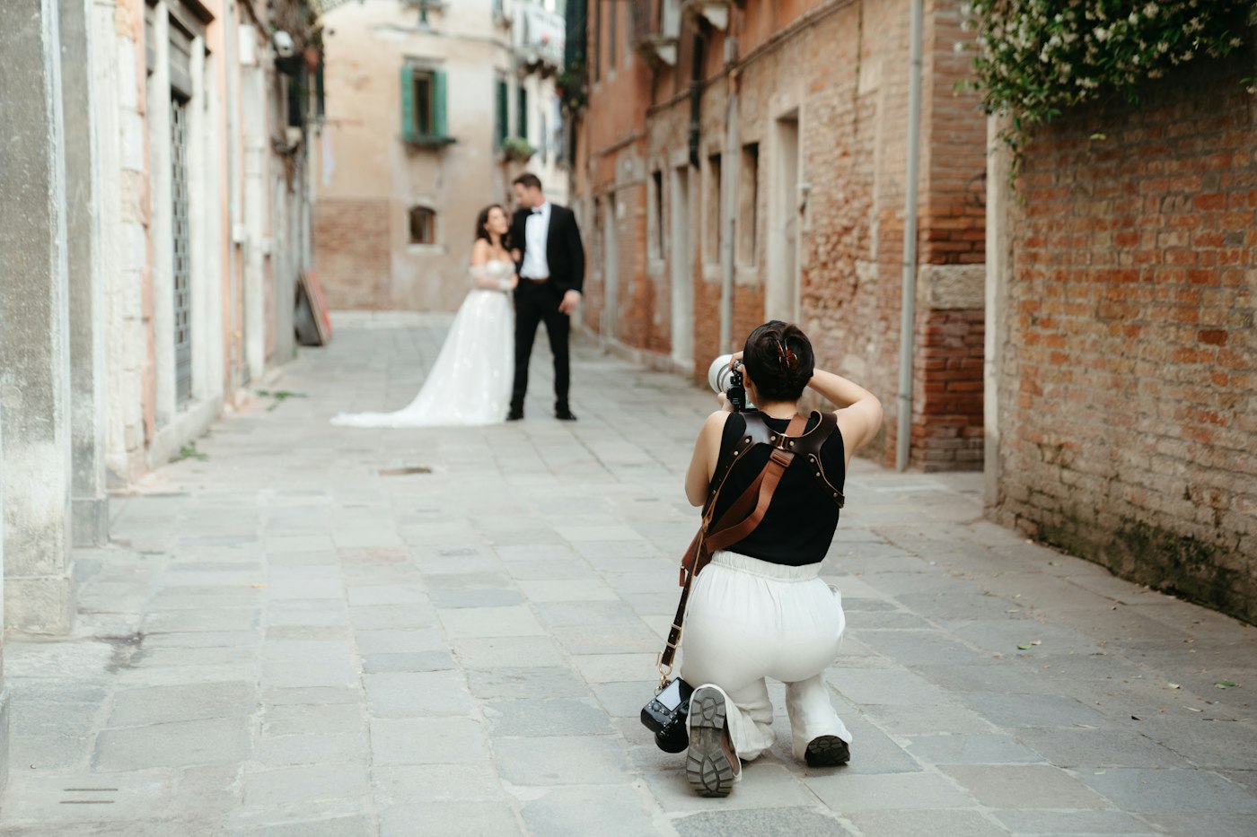 Eine Hochzeitsfotografin kniet auf dem Pflaster einer schmalen Gasse in Venedig und fotografiert das Brautpaar weiter hinten