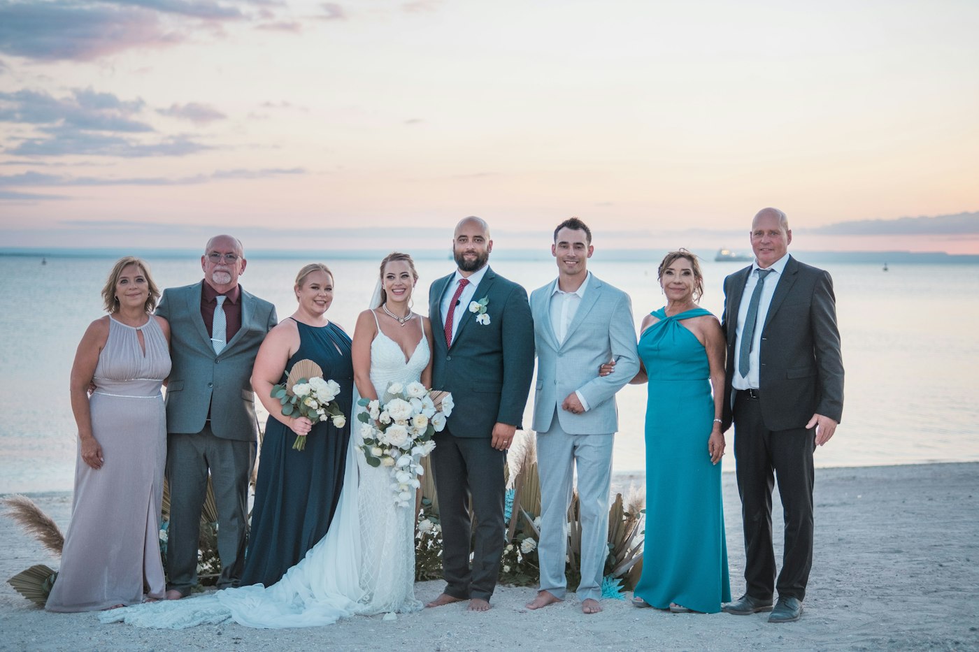 Ein Brautpaar am Strand bei Sonnenuntergang mit sechs Familienmitgliedern in festlicher Kleidung, die Braut hält einen weißen Strauß