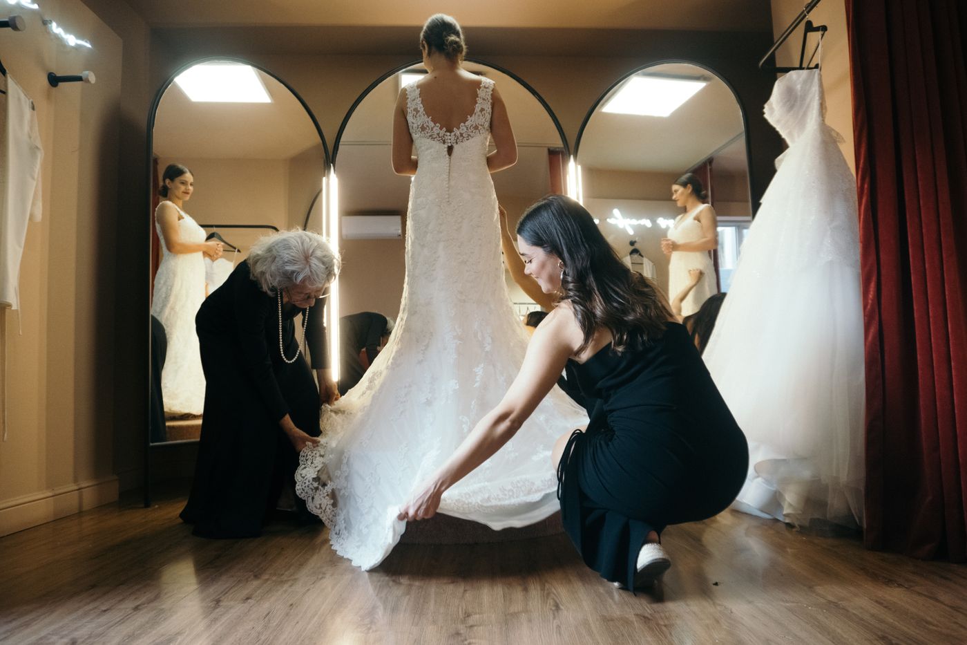 Eine Braut im Spitzenkleid steht in einer Umkleide vor gewölbten Spiegeln, während zwei Frauen die Schleppe glatt ziehen