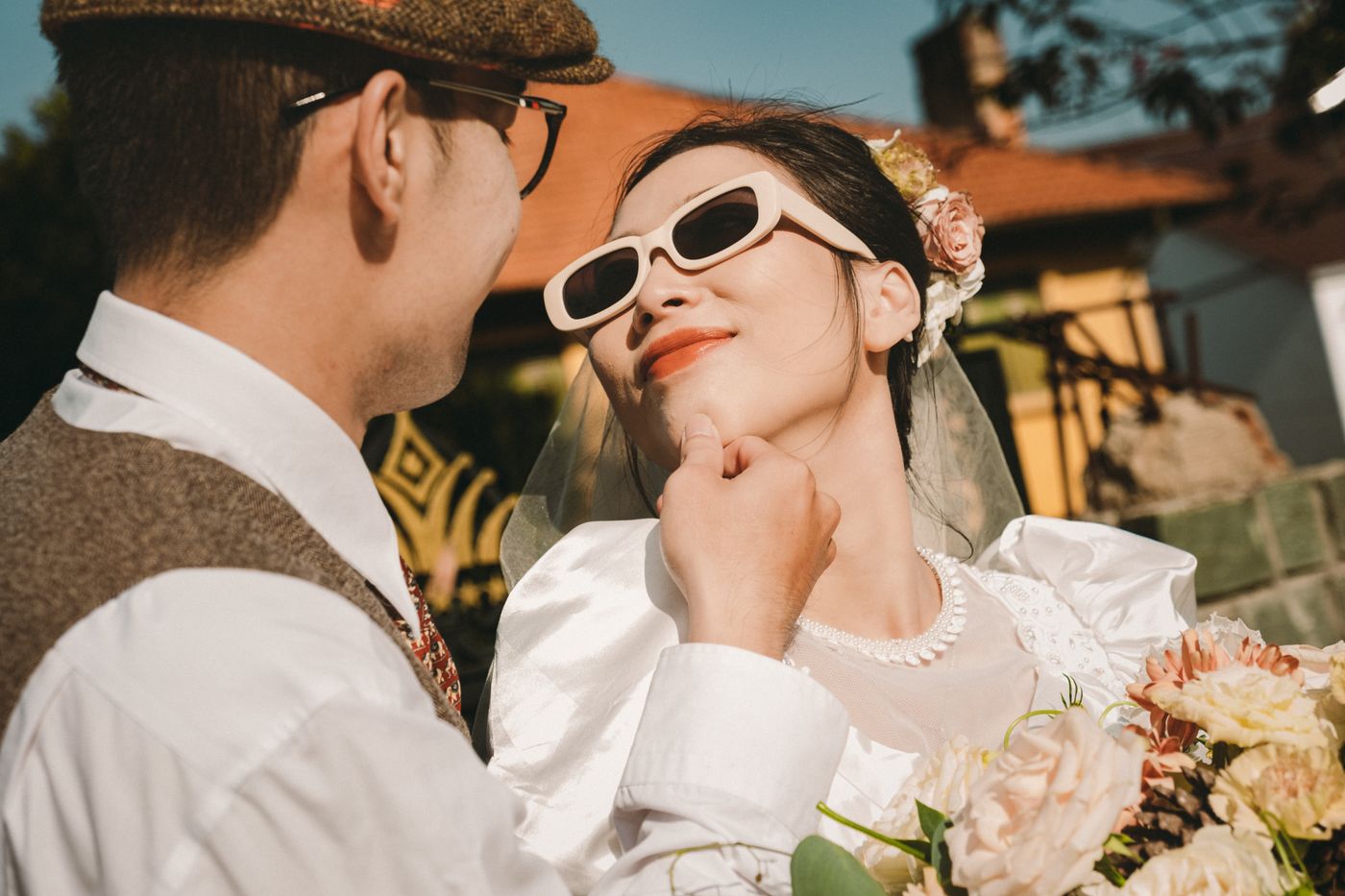 Una pareja con estilo retro: él con gorra plana y chaleco de tweed, ella de raso con perlas, velo y gafas de sol blancas de ojo de gato