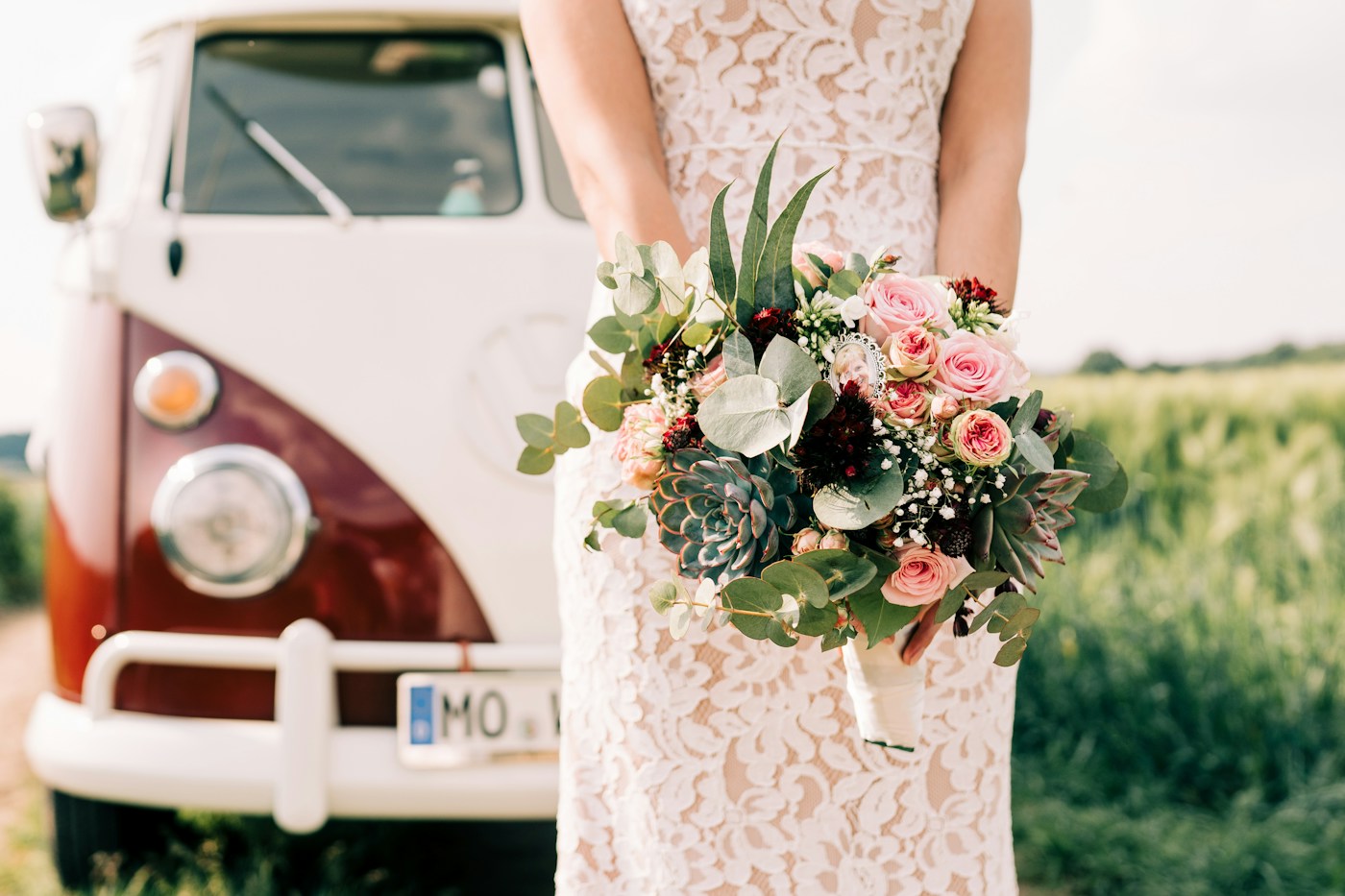 Una novia con un vestido ajustado de encaje sostiene un ramo de rosas rosas y suculentas junto a una furgoneta Volkswagen antigua de color crema y rojo