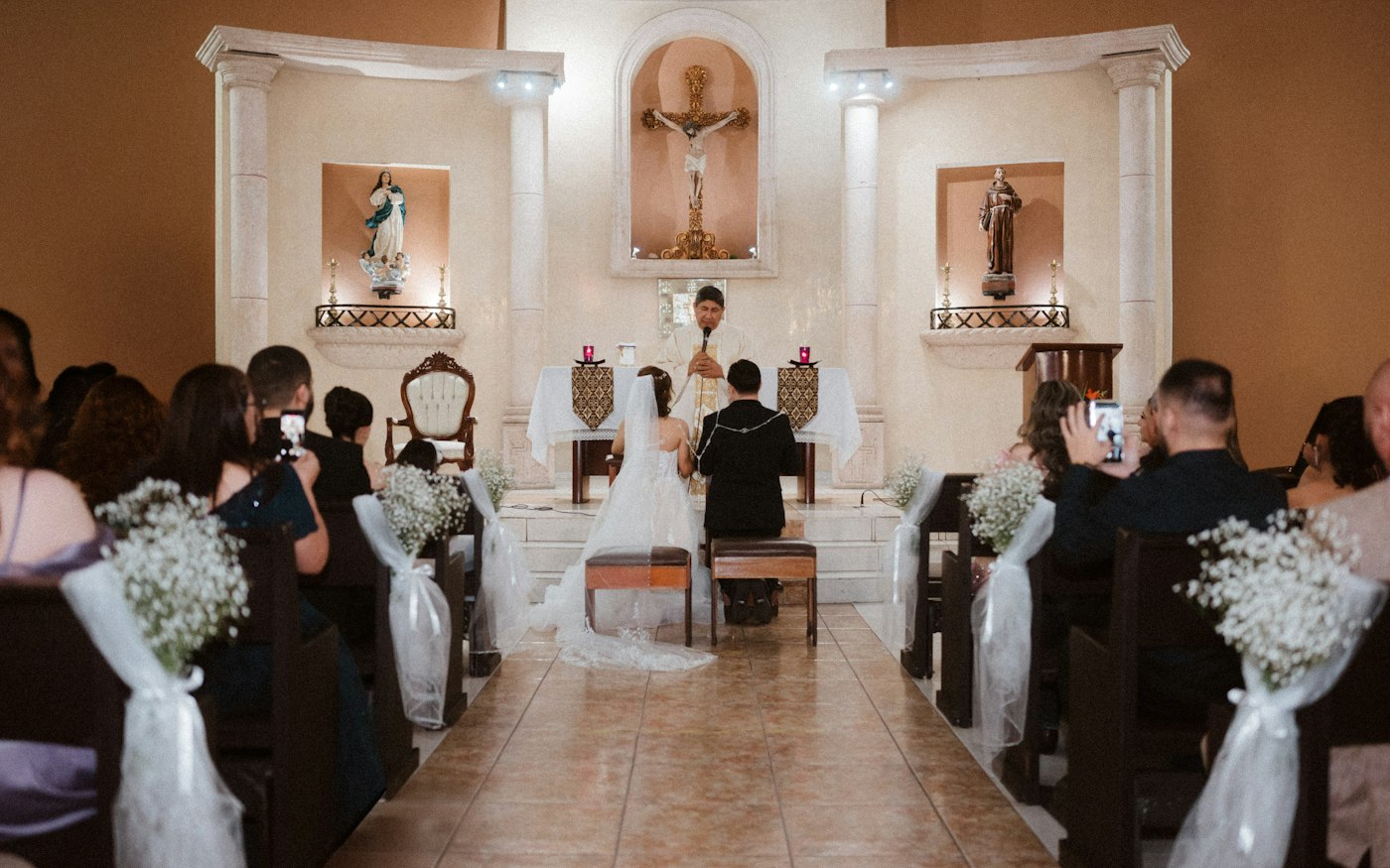 Kirchenbänke mit weißen Tüllschleifen und Sträußchen aus Schleierkraut, vorne kniet das Brautpaar am Altar
