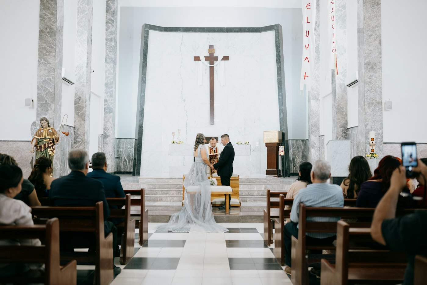 Ein Brautpaar hält sich am Altar einer hellen Kirche mit Marmorwänden an den Händen, die Gäste schauen aus den Bänken zu
