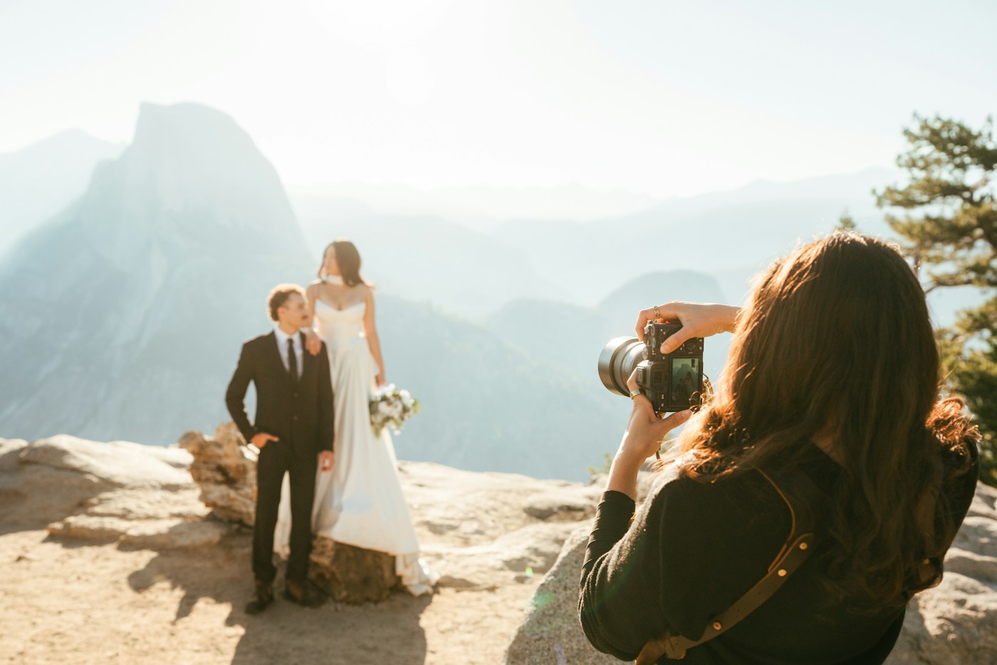 Una fotógrafa con cámara retrata a una pareja de novios en un mirador rocoso con montañas de granito al fondo
