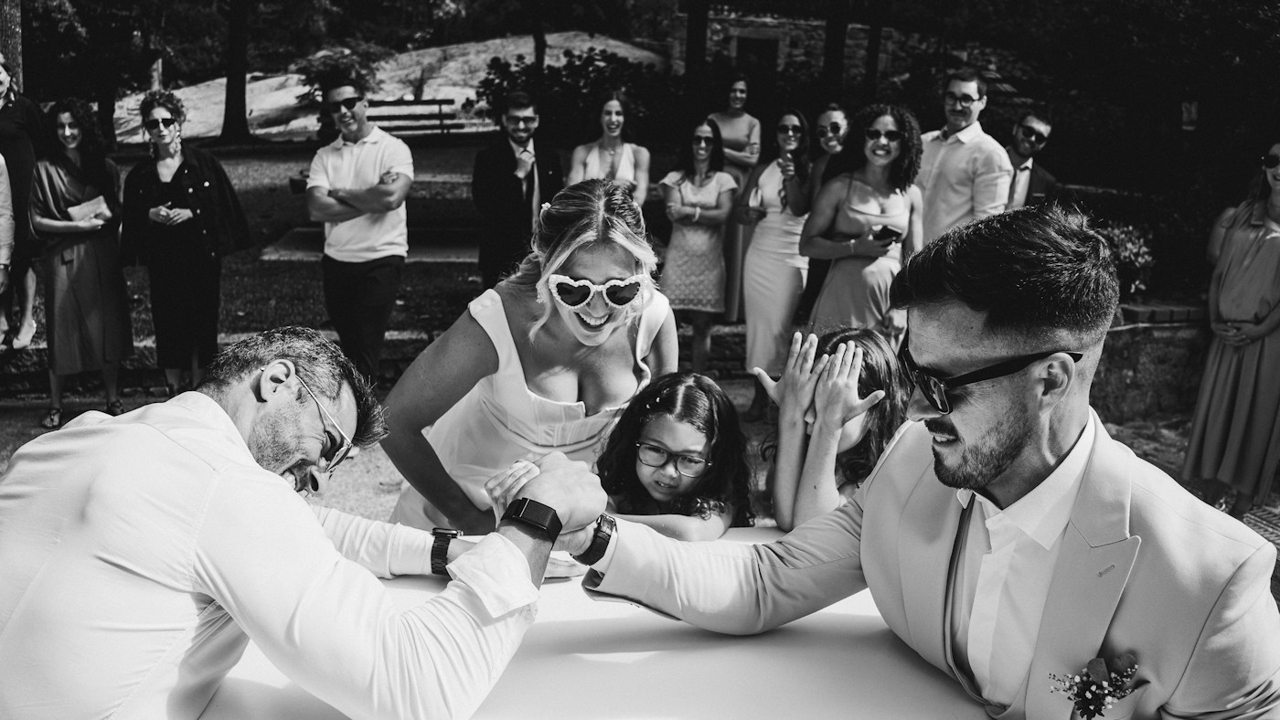 Una novia con gafas de sol en forma de corazón se ríe mientras dos hombres echan un pulso en una mesa y los invitados miran en un jardín