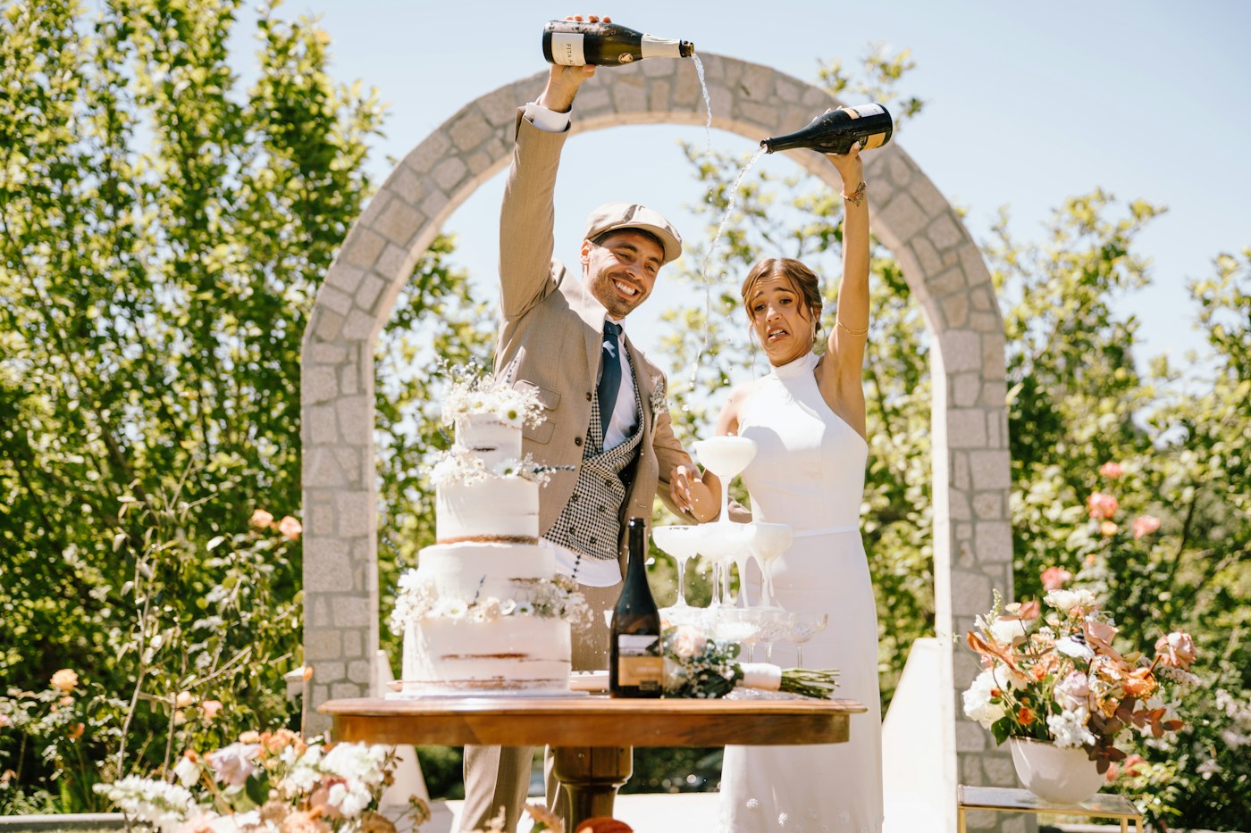 Los novios, entre risas, sirven cava en una torre de copas junto a una tarta desnuda de tres pisos bajo un arco de piedra en un jardín soleado