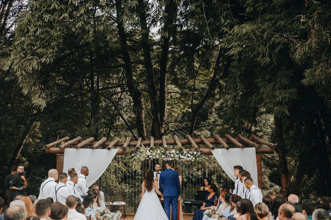 Ein Brautpaar heiratet unter einer Holzpergola mit weißen Tüchern in einem bewaldeten Garten, die Gäste sitzen links und rechts vom Gang