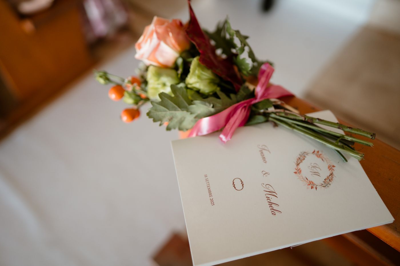 Un librito doblado de color crema con los nombres de los novios y una corona de hojas descansa sobre un banco de iglesia bajo un ramito de rosas atado con una cinta rosa