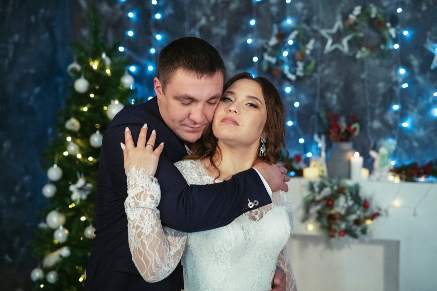 Brautpaar umarmt sich vor einem geschmückten Weihnachtsbaum mit blauer Lichterkette und einem kleinen Tisch mit Kerzen