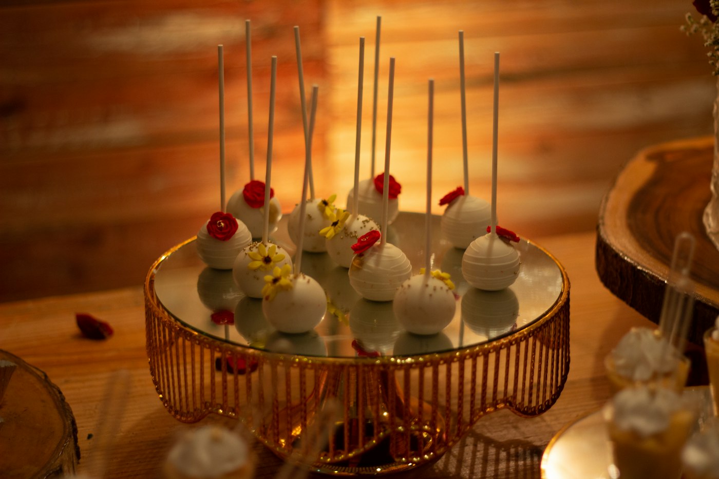 Weiß überzogene Cake Pops mit kleinen Zuckerblüten auf einer goldenen Etagere mit Spiegelplatte an einem Desserttisch