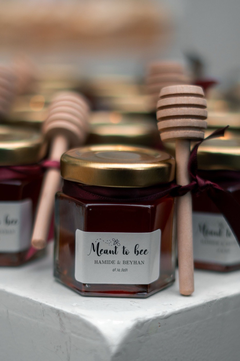 Small hexagonal honey jars with gold lids, wooden honey dippers and 'Meant to bee' labels with the couple's names, set out as wedding favours